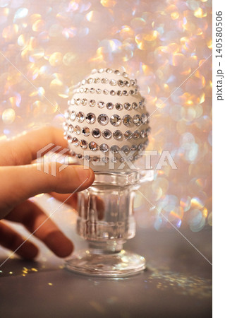 Decorative white egg adorned with rhinestones held by a hand, placed on a clear glass stand against a shimmering bokeh background Decorative white egg adorned with rhinestones held by a hand, placed on a clear glass stand against a shimmering bokeh background 140580506