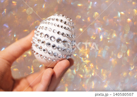 Decorative white Easter egg embellished with rhinestones held in a hand against a shimmering, blurred background of soft pastel colors Decorative white Easter egg embellished with rhinestones held in a hand against a shimmering, blurred background of soft pastel colors 140580507