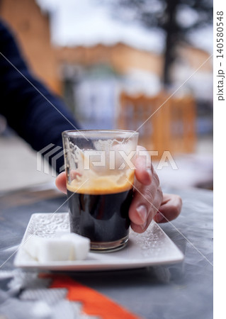 Close-up of a hand holding a glass of espresso on a white plate with sugar cubes, set against a blurred outdoor cafe background, capturing a moment of coffee enjoyment Close-up of a hand holding a glass of espresso on a white plate with sugar cubes, set against a blurred outdoor cafe background, capturing a moment of coffee enjoyment 140580548