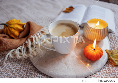 Cozy autumn scene featuring a white coffee cup on a marble tray, surrounded by decorative pumpkins, a candle, and a book, creating a warm and inviting atmosphere 140580555