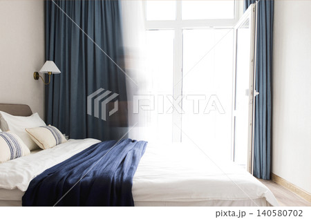 Modern bedroom interior with a neatly made bed, decorative pillows, and blue curtains, featuring a bright window and a soft blanket draped across the bed Modern bedroom interior with a neatly made bed, decorative pillows, and blue curtains, featuring a bright window and a soft blanket draped across the bed 140580702