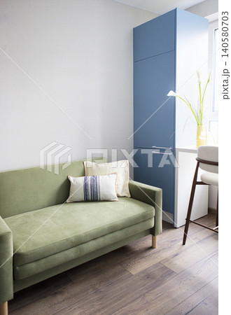 Green sofa with decorative pillows positioned beside a blue cabinet and a window, featuring a plant and a wooden chair in a modern interior setting Green sofa with decorative pillows positioned beside a blue cabinet and a window, featuring a plant and a wooden chair in a modern interior setting 140580703