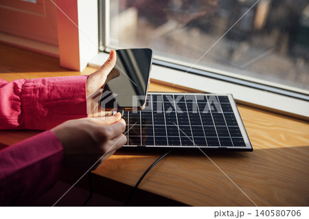 Person in pink shirt connects smartphone to small solar panel on wooden windowsill, with sunlight illuminating the scene and a blurred outdoor view in the background Person in pink shirt connects smartphone to small solar panel on wooden windowsill, with sunlight illuminating the scene and a blurred outdoor view in the background 140580706