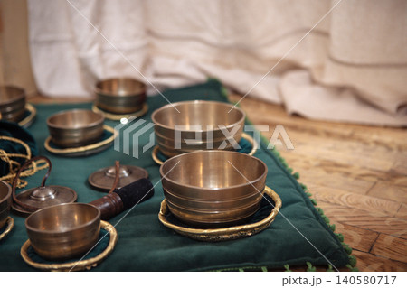 Set of brass singing bowls arranged on a green fabric mat with decorative edges, wooden mallet and additional bowls visible in a serene indoor setting Set of brass singing bowls arranged on a green fabric mat with decorative edges, wooden mallet and additional bowls visible in a serene indoor setting 140580717