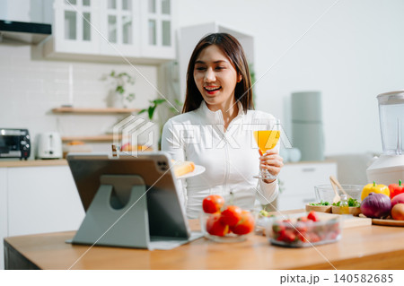 Healthy woman cooking at home while video calling online. Clean eating, nutrition, fresh vegetables, smoothie and digital lifestyle concept in modern kitchen. Healthy woman cooking at home while video calling online. Clean eating, nutrition, fresh vegetables, smoothie and digital lifestyle concept in modern kitchen. 140582685