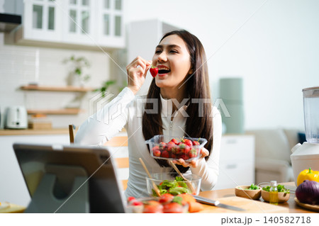 Young woman eating fresh salad in modern kitchen while using tablet. Healthy food, diet lifestyle, online cooking tutorial, wellness and clean eating concept. 140582718