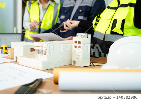 Engineer team in safety helmets discussing house model and blueprint. Construction planning, architecture design, project management Engineer team in safety helmets discussing house model and blueprint. Construction planning, architecture design, project management 140582986