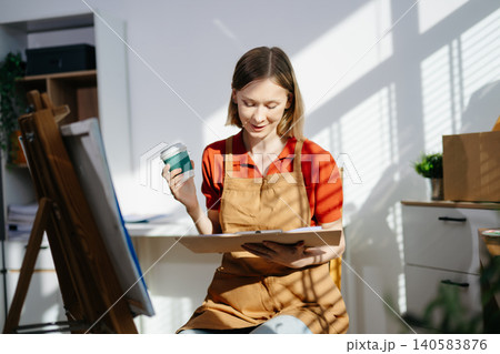 A focused female artist holding her coffee while sketching ideas in her sunlit studio, capturing creativity, peaceful workflow, and an inspiring artistic routine. 140583876
