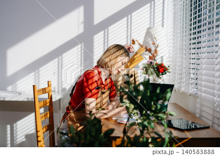 A focused female artist painting at a wooden desk in a bright home studio with art tools, laptop, and natural sunlight 140583888