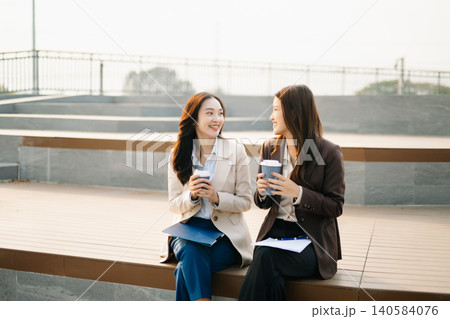 Asian businesswomen reviewing documents and writing notes outdoors. Modern teamwork, planning, and professional collaboration in a casual corporate lifestyle setting. Asian businesswomen reviewing documents and writing notes outdoors. Modern teamwork, planning, and professional collaboration in a casual corporate lifestyle setting. 140584076
