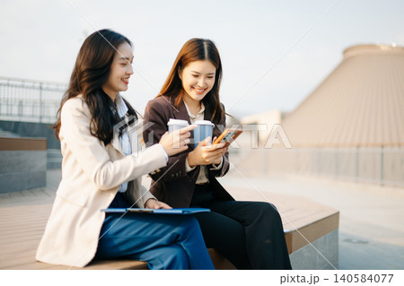 Asian businesswomen reviewing documents and writing notes outdoors. Modern teamwork, planning, and professional collaboration in a casual corporate lifestyle setting. Asian businesswomen reviewing documents and writing notes outdoors. Modern teamwork, planning, and professional collaboration in a casual corporate lifestyle setting. 140584077