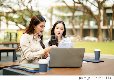 Two businesswomen working together with laptop and documents in outdoor workspace, representing teamwork, startup collaboration, digital business planning Two businesswomen working together with laptop and documents in outdoor workspace, representing teamwork, startup collaboration, digital business planning 140584113