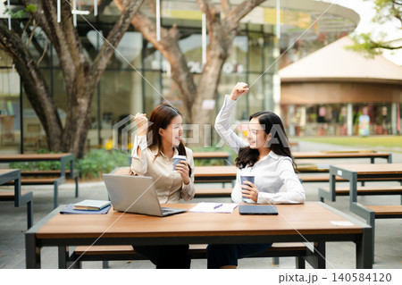 Two businesswomen celebrating success during an outdoor coffee meeting with laptop and documents, representing teamwork, startup collaboration Two businesswomen celebrating success during an outdoor coffee meeting with laptop and documents, representing teamwork, startup collaboration 140584120