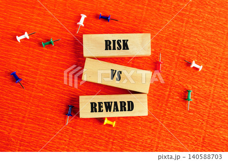 Blocks with the words risk and reward sit on an orange surface with colorful push pins around them. This setup captures a contrast between two concepts 140588703