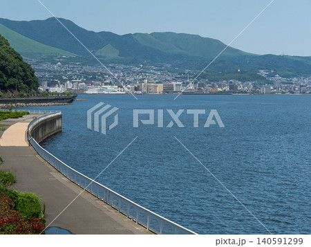 Scenic view of a waterfront promenade along the blue sea with a coastal city and mountains in the background. Scenic view of a waterfront promenade along the blue sea with a coastal city and mountains in the background. 140591299