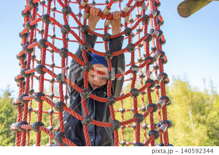 A young boy in a blue beanie carefully climbing up a red rope cargo net at an outdoor adventure playground. Concept of childhood bravery, active lifestyle, and summer vacation. Active child playing on A young boy in a blue beanie carefully climbing up a red rope cargo net at an outdoor adventure playground. Concept of childhood bravery, active lifestyle, and summer vacation. Active child playing on 140592344