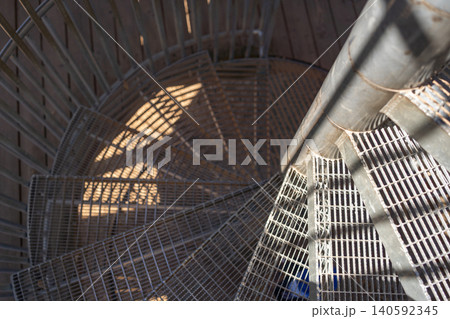 Dramatic high contrast top down shot of an outdoor metal spiral staircase. Bright sunlight creates strong graphic shadows on the steel grid steps, emphasizing industrial architecture. Looking down a 140592345