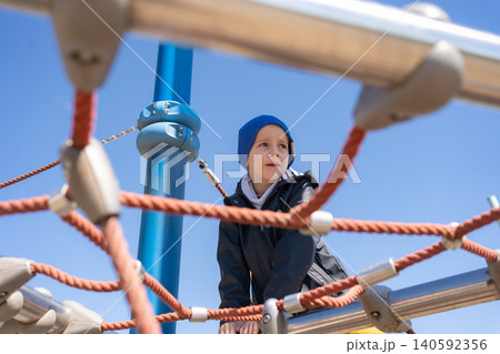 A young boy wearing a blue beanie framed by playground ropes at an outdoor adventure park. Candid lifestyle portrait capturing child concentration, outdoor activity, and active childhood. Cute little A young boy wearing a blue beanie framed by playground ropes at an outdoor adventure park. Candid lifestyle portrait capturing child concentration, outdoor activity, and active childhood. Cute little 140592356
