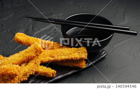 Crispy food served with chopsticks and a bowl of sauce on a dark surface 140594546