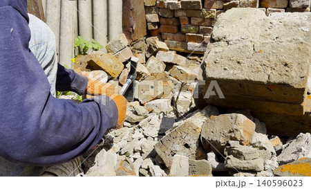 Male hands in protective gloves uses hammer to remove old cement from used bricks during renovation work. The process involves carefully cleaning brick surface to prepare it for reuse in construction. Male hands in protective gloves uses hammer to remove old cement from used bricks during renovation work. The process involves carefully cleaning brick surface to prepare it for reuse in construction. 140596023