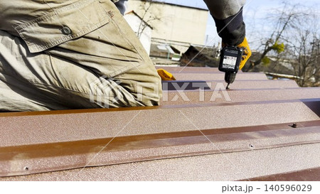 Male hands in gloves use cordless screwdriver to fasten roofing sheets in house roof. Young worker secures screws into surface performing careful installation work during residential construction 140596029