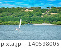 White sailboat sailing on a wide river with lush green hills under blue sky 140598005