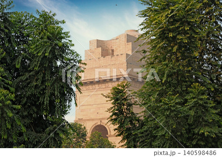 Delhi, India, November 28, 2025: The historic India Gate monument made of sandstone with India inscription viewed through a natural frame of green tree leaves under a blue sky. Delhi, India, November 28, 2025: The historic India Gate monument made of sandstone with India inscription viewed through a natural frame of green tree leaves under a blue sky. 140598486