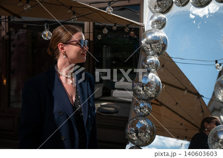 Smiling young woman in sunglasses stands beside mirror framed with disco balls, creating sparkling reflections and stylish contemporary cafe atmosphere. Lifestyle, creative lighting, fashion Smiling young woman in sunglasses stands beside mirror framed with disco balls, creating sparkling reflections and stylish contemporary cafe atmosphere. Lifestyle, creative lighting, fashion 140603603
