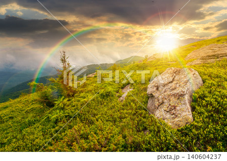 sunset over green landscape of mountain in summer. view of rock among grass on the hillside. travel background under a cloudy sky. mountain scenery with rolling hills. weather with rainbow 140604237