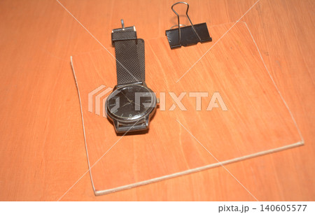 Black analog wristwatch and a single metal binder on a wooden desk with fastener 140605577