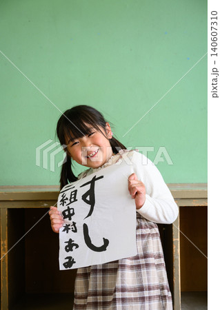 教室の後ろで笑顔で「すし」の習字作品を見せる女子小学生の写真 140607310