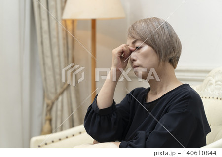 リビングのソファで体調不良で頭をかかえるシニア女性 リビングのソファで体調不良で頭をかかえるシニア女性 140610844