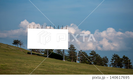 A large blank billboard stands on metal supports above a green sloping hill, with pine trees in the background and soft white clouds across a clear blue sky A large blank billboard stands on metal supports above a green sloping hill, with pine trees in the background and soft white clouds across a clear blue sky 140617695