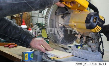 A workflow in a woodworking shop: a craftsman making a cut on a miter saw. The man's right hand rests on the saw handle, controlling the blade's downward movement. His left hand presses the wood A workflow in a woodworking shop: a craftsman making a cut on a miter saw. The man's right hand rests on the saw handle, controlling the blade's downward movement. His left hand presses the wood 140618711