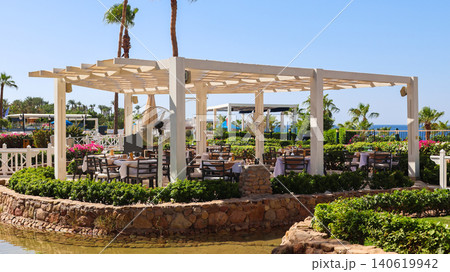 Cozy summer cafe surrounded by lush greenery and a serene pond, perfect for relaxation and outdoor dining 140619942