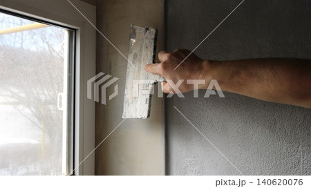 Interior finishing work, leveling a window slope. A man's hand holds a wide facade spatula and presses the blade of the spatula against the plane of the slope to the left of the wall corner Interior finishing work, leveling a window slope. A man's hand holds a wide facade spatula and presses the blade of the spatula against the plane of the slope to the left of the wall corner 140620076
