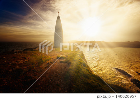 A conical tower stands on a grassy cliff overlooking the ocean during a bright sunset with sunbeams shining on the water. A conical tower stands on a grassy cliff overlooking the ocean during a bright sunset with sunbeams shining on the water. 140620957