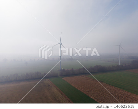 Aerial view of industrial wind turbines standing in misty green and brown agricultural fields under bright morning sun fog in the Dutch countryside, Netherlands. Aerial view of industrial wind turbines standing in misty green and brown agricultural fields under bright morning sun fog in the Dutch countryside, Netherlands. 140622619