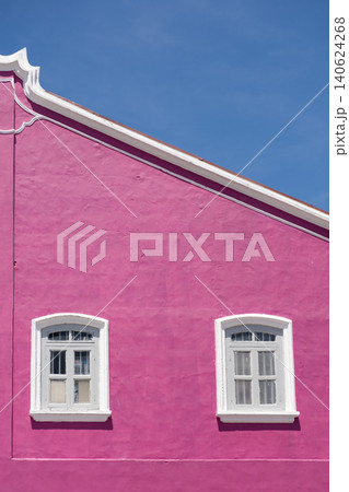 Bright pink wall of a building featuring two white-framed windows under a clear blue sky, showcasing vibrant color contrast and architectural details in a sunny environment Bright pink wall of a building featuring two white-framed windows under a clear blue sky, showcasing vibrant color contrast and architectural details in a sunny environment 140624268