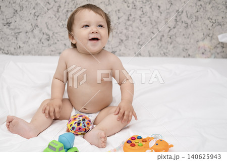 Adorable diaper baby boy sitting on white bed and reaching for toy Adorable diaper baby boy sitting on white bed and reaching for toy 140625943