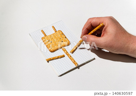 Hand arranging pretzel sticks and a square cracker on a graph paper notebook Hand arranging pretzel sticks and a square cracker on a graph paper notebook 140630597