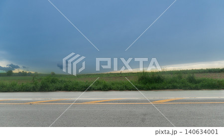 Horizontal or Landscape showing an empty wet rainy asphalt roadway. Beneath a wide dark storm cloud formation. Lush green grassy fields border the modern concrete landscape layout. 140634001