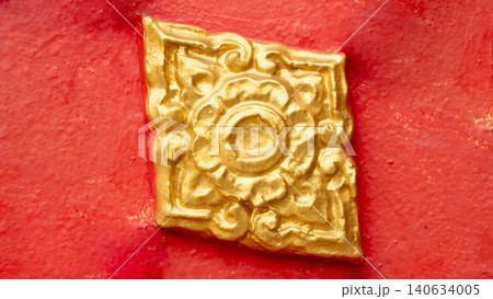 Close-up view showing a single square golden flower-shaped Lai Kanok. Thai ornament ornament attached firmly to a rough-textured red stucco wall panel background. Close-up view showing a single square golden flower-shaped Lai Kanok. Thai ornament ornament attached firmly to a rough-textured red stucco wall panel background. 140634005