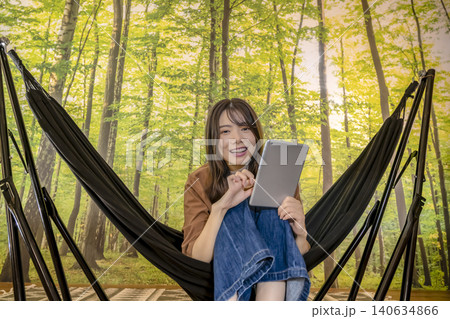 キャンプでハンモックに座ってタブレットを操作する若い女性 140634866