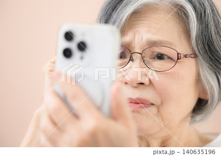 ピンク背景で老眼鏡でスマホを見るグレイヘアのシニア女性 文字が見えにくい詐欺かな?と疑う悩むイメージ ピンク背景で老眼鏡でスマホを見るグレイヘアのシニア女性 文字が見えにくい詐欺かな?と疑う悩むイメージ 140635196