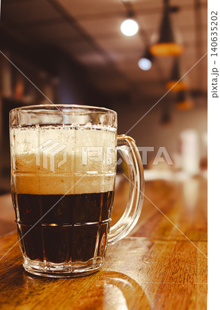Dark stout beer filling a transparent glass mug, showing a rich white foam head. Pub in the blurred background Dark stout beer filling a transparent glass mug, showing a rich white foam head. Pub in the blurred background 140635202
