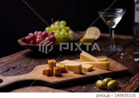 Still life displaying gourmet appetizers including cheeses, grapes, olives, and a drink with lemon on a rustic wooden table Still life displaying gourmet appetizers including cheeses, grapes, olives, and a drink with lemon on a rustic wooden table 140635210