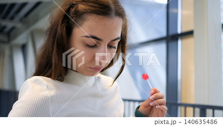 Close portrait of young woman near window holding lollipop with thoughtful expression, soft daylight illuminating face and textured sweater, intimate quiet mood in transit corridor. Close portrait of young woman near window holding lollipop with thoughtful expression, soft daylight illuminating face and textured sweater, intimate quiet mood in transit corridor. 140635486