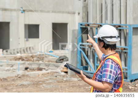 Construction engineer wearing safety helmet reflective vest inspecting building project tablet while pointing at development progress site Construction engineer wearing safety helmet reflective vest inspecting building project tablet while pointing at development progress site 140635655