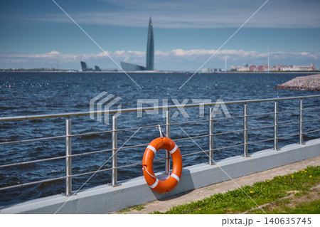 An orange-colored lifebuoy hangs on the embankment fence in sunny weather, finland gulf, with the sea and a skyscraper in the background. An orange-colored lifebuoy hangs on the embankment fence in sunny weather, finland gulf, with the sea and a skyscraper in the background. 140635745